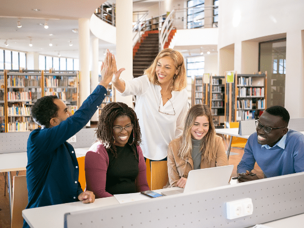 Students learning in a library. One student is high-fiving the teacher.