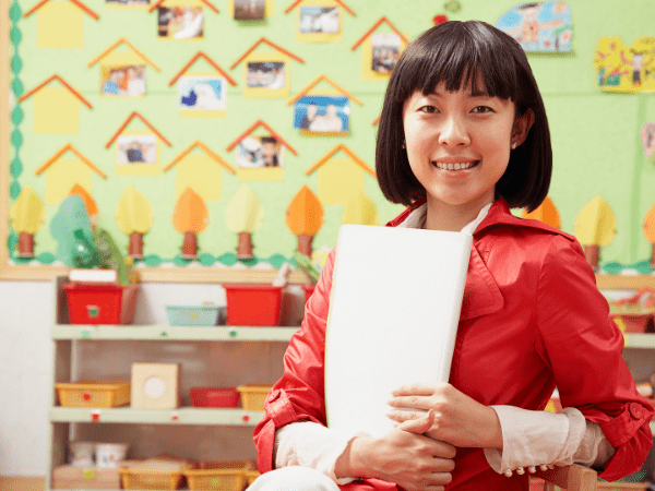 A teacher smiling in her classroom holding a pile of worksheets.